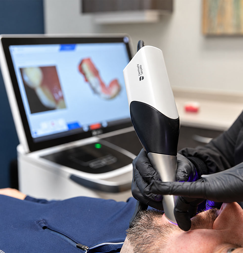 Dental professional using an intraoral scanner on a patient, with a digital model of teeth on a nearby monitor.