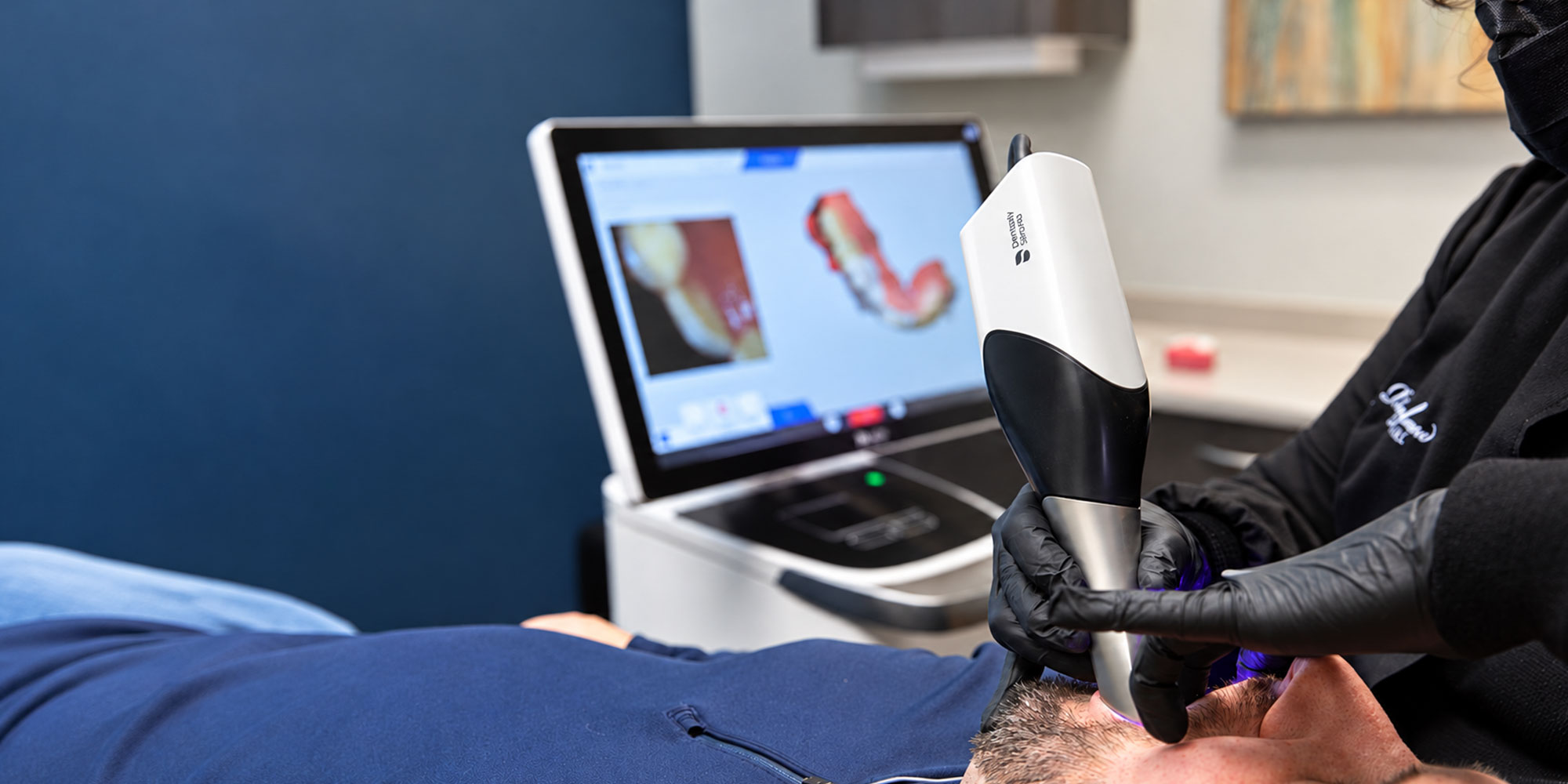 Dental professional using an intraoral scanner on a patient, with a digital model of teeth displayed on a nearby monitor.