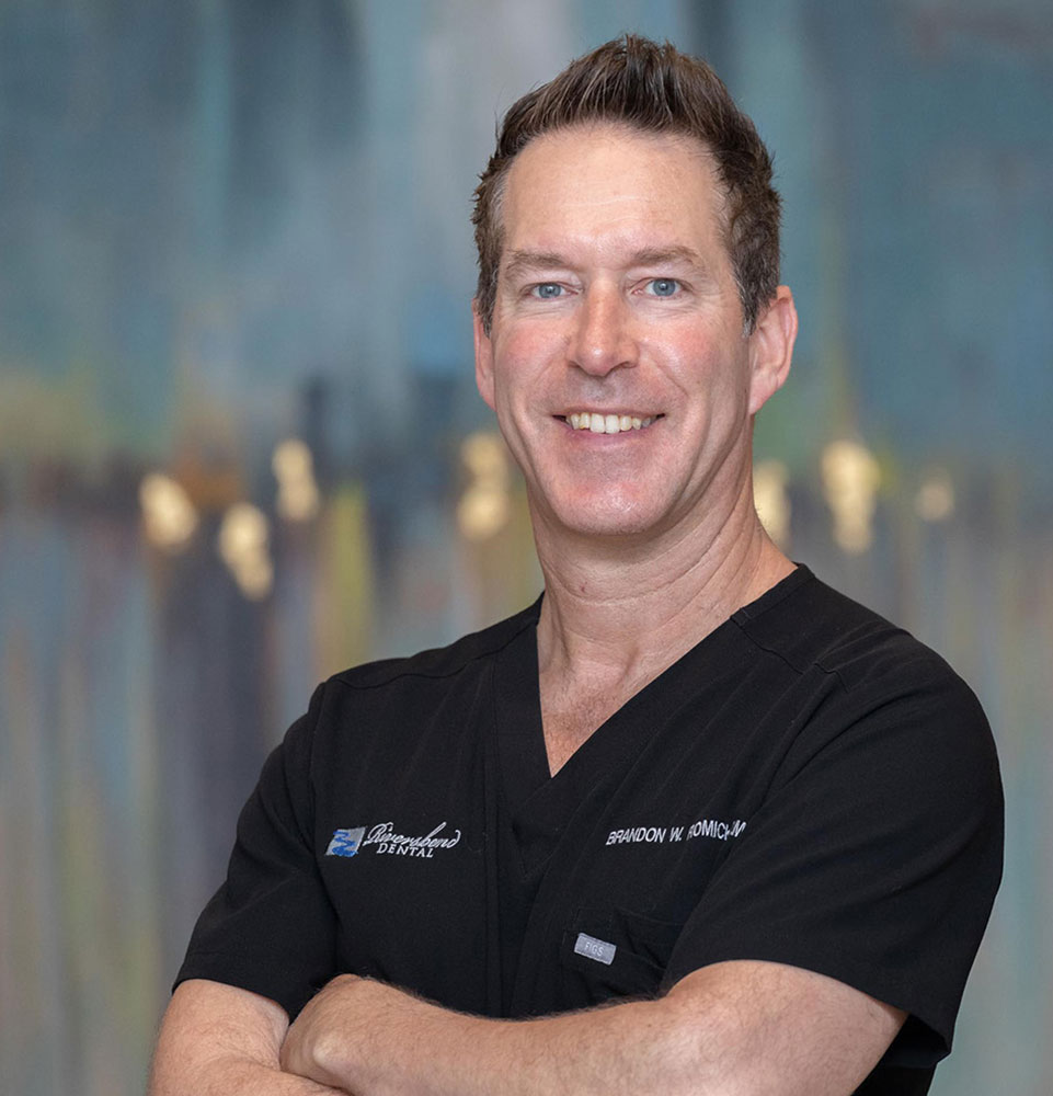 Dr. Brandon Romick smiling with his arms crossed, wearing black dental scrubs.