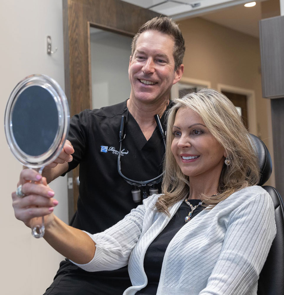 Smiling patient looking into a handheld mirror with Dr. Brandon Romick and a dental team member beside her.
